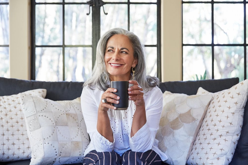 An older woman sitting on a couch holding a coffee mug and smiling.