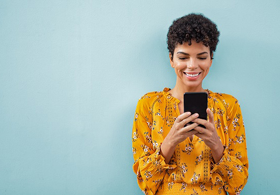 Smiling black woman using smart phone and standing against a light blue wall.