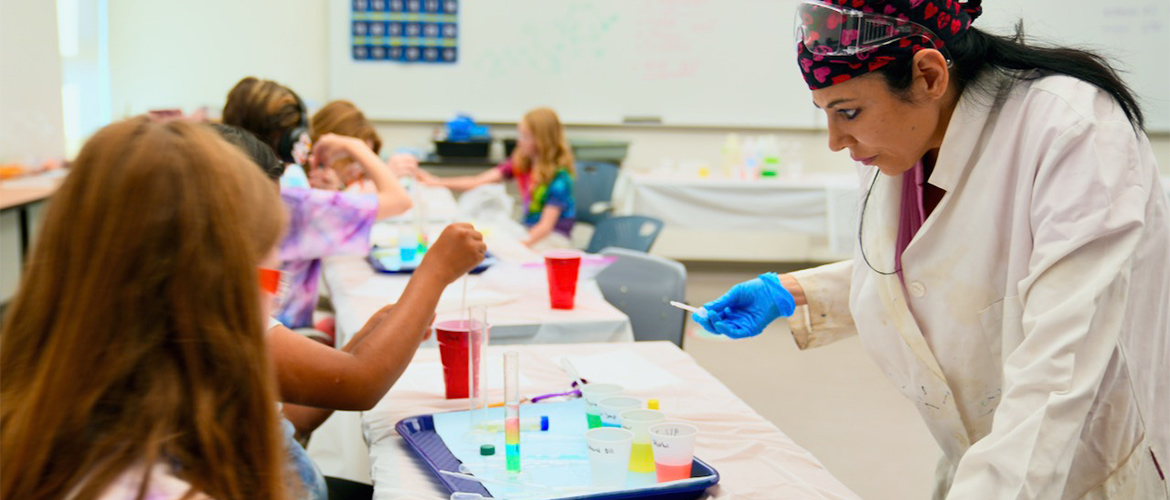 A woman mentor in a white lab coat leans over a table to help young girls mixing solutions in a cylinder. 