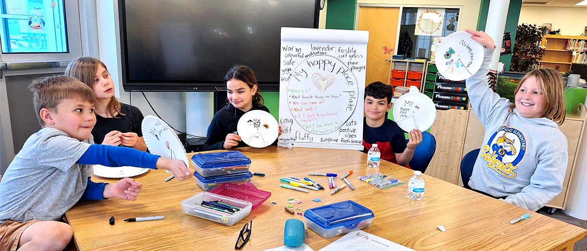 Children sitting around a table hold up art projects