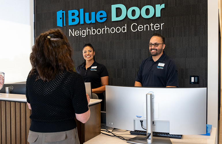 two individuals smile behind counter greeting a woman